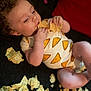Wynter is registered to the contest to win money with this photo: baby, child, tortilla_chip, snack, curly_hair, onesie, food, eating, lying_down, infant, cute, hand, face, skin, relaxing, closeup, indoor, soft_lighting, casual, portrait
