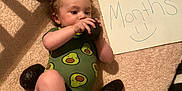 Wynter joined the competition — help win amazing prizes! baby, infant, avocado, onesie, crib, blanket, milestone, age_sign, handwritten_sign, cuddly, cute, smiling_face, food, fruit, playful, soft_texture, indoor, child, person, lying_down