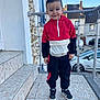 black_pants, boy, car, casual_clothing, child, daylight, happy, house, outdoor, porch, railing, red_jacket, residential_area, short_hair, sidewalk, smiling, sneakers, standing, tiles, young