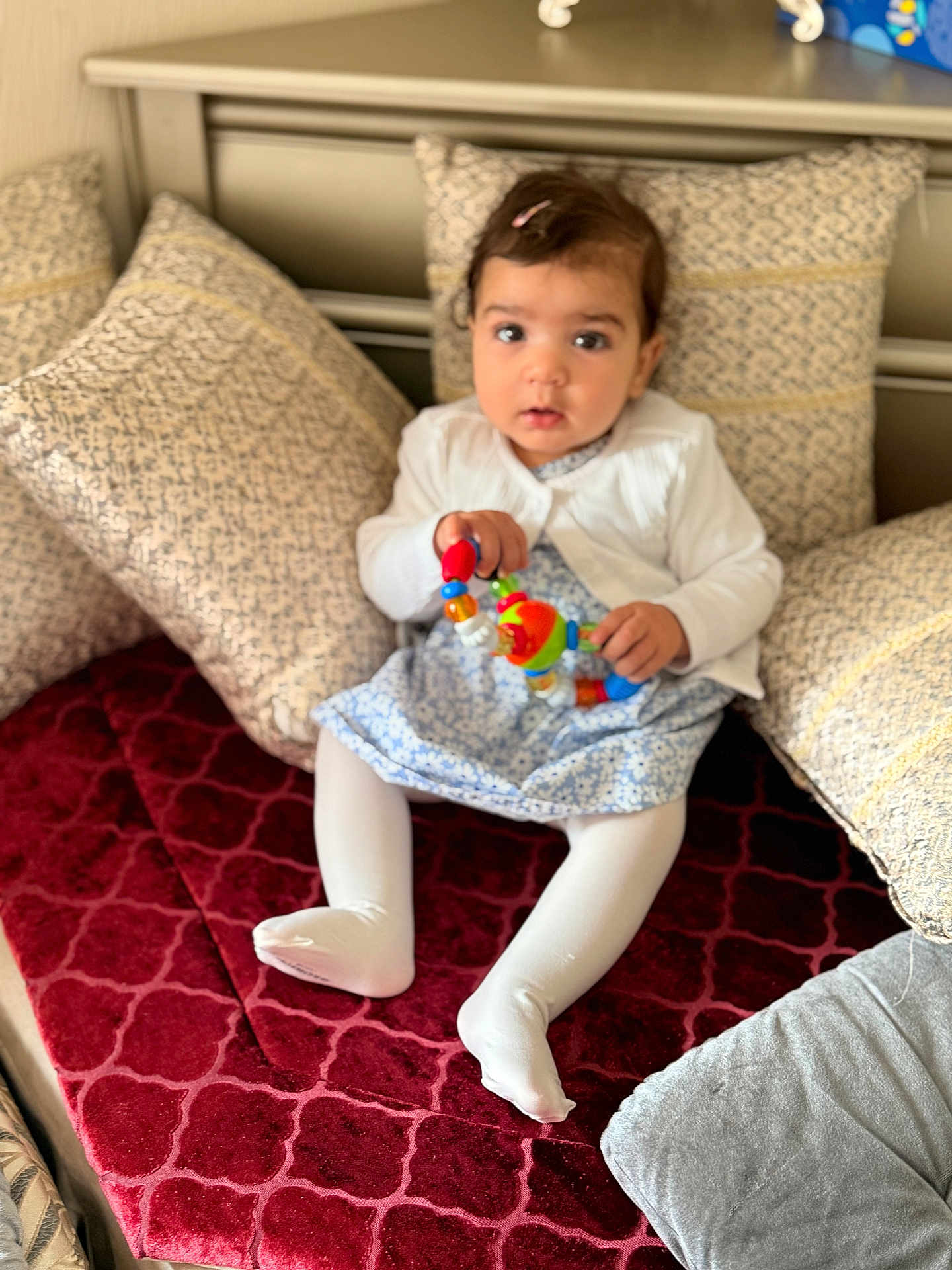 Omnia participe au concours pour gagner de l'argent avec cette photo : toddler, child, toy, pillow, red_velvet, couch, white_tights, blue_dress, hair_clip, indoor, plush, patterned_pillows, seated, curious_expression, baby, person, furniture, soft_lighting, home, portrait