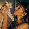 woman, cat, orange_tabby, tattoo, earrings, portrait, indoor, warm_lighting, holding, pets, expression, face, animal, human, intimate_moment, closeup, hair, skin, ear, eyes