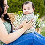 Giovanne is registered to the contest to win money with this photo: baby, woman, outdoor, greenery, flowers, holding, child, portrait, nature, casual_clothing, jeans, green_top, pouting, cute, tender, mother_and_child, garden, daylight, smile, affection