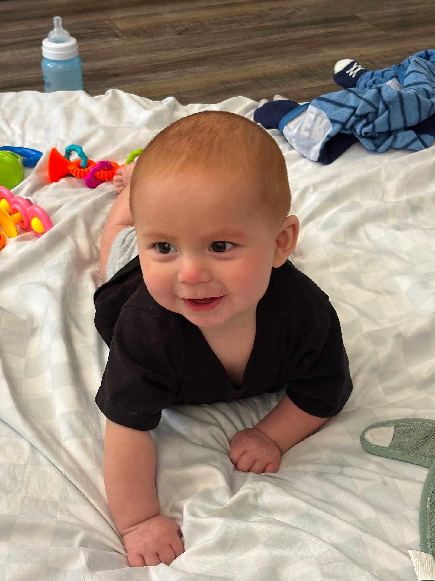 Nathaniel joined the competition — help win amazing prizes! baby, crawling, black_shirt, blanket, toy, bottle, floor, indoor, smiling, child, infant, person, cute, happy, head, hand, face, clothing, wooden_floor, playtime
