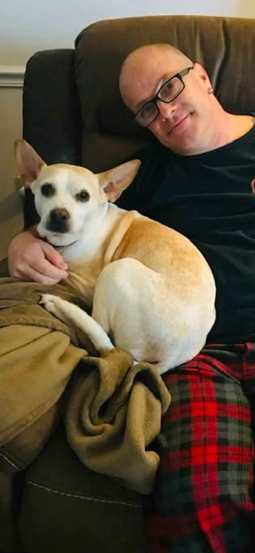 Lucy is registered to the contest to win money with this photo: man, glasses, dog, couch, blanket, plaid_pajamas, pet, sitting, living_room, recliner, paw, ear, fur, smile, human, male, hand, cozy, companion, home_interior