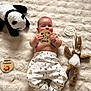 baby, infant, plush_toy, panda, bunny, blanket, card, text, clothing, socks, cute, lying_down, child, soft_texture, patterned_pants, handheld_object, indoors, portrait, smiling, young_child
