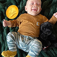 Woodson is registered to the contest to win money with this photo: baby, infant, smiling, blanket, plush_toy, black_dog_toy, brown_shirt, camo_pants, mustard_socks, wooden_plaque, text, 2_months, lying_down, happy, cute, child, indoors, portrait, soft_texture, cozy