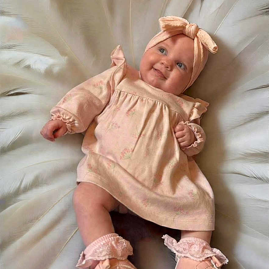 Ayla-Louise is registered to the contest to win money with this photo: apparel, baby, blanket, child, cute, dress, fashion, feathers, footwear, happy, headband, indoors, infant, lying_down, person, portrait, shoes, smiling, soft_colors, toddler