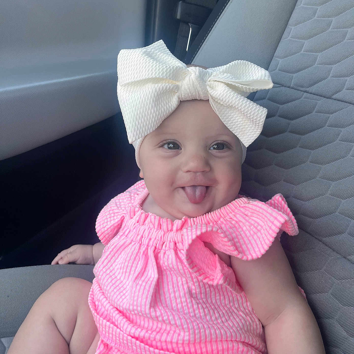Kaislee is registered to the contest to win money with this photo: baby, car_interior, car_seat, child, chubby_arms, clothing, cute, happy, headwear, indoor, infant, person, pink_outfit, portrait, seat, seatbelt, sitting, smiling, tongue_out, white_bow