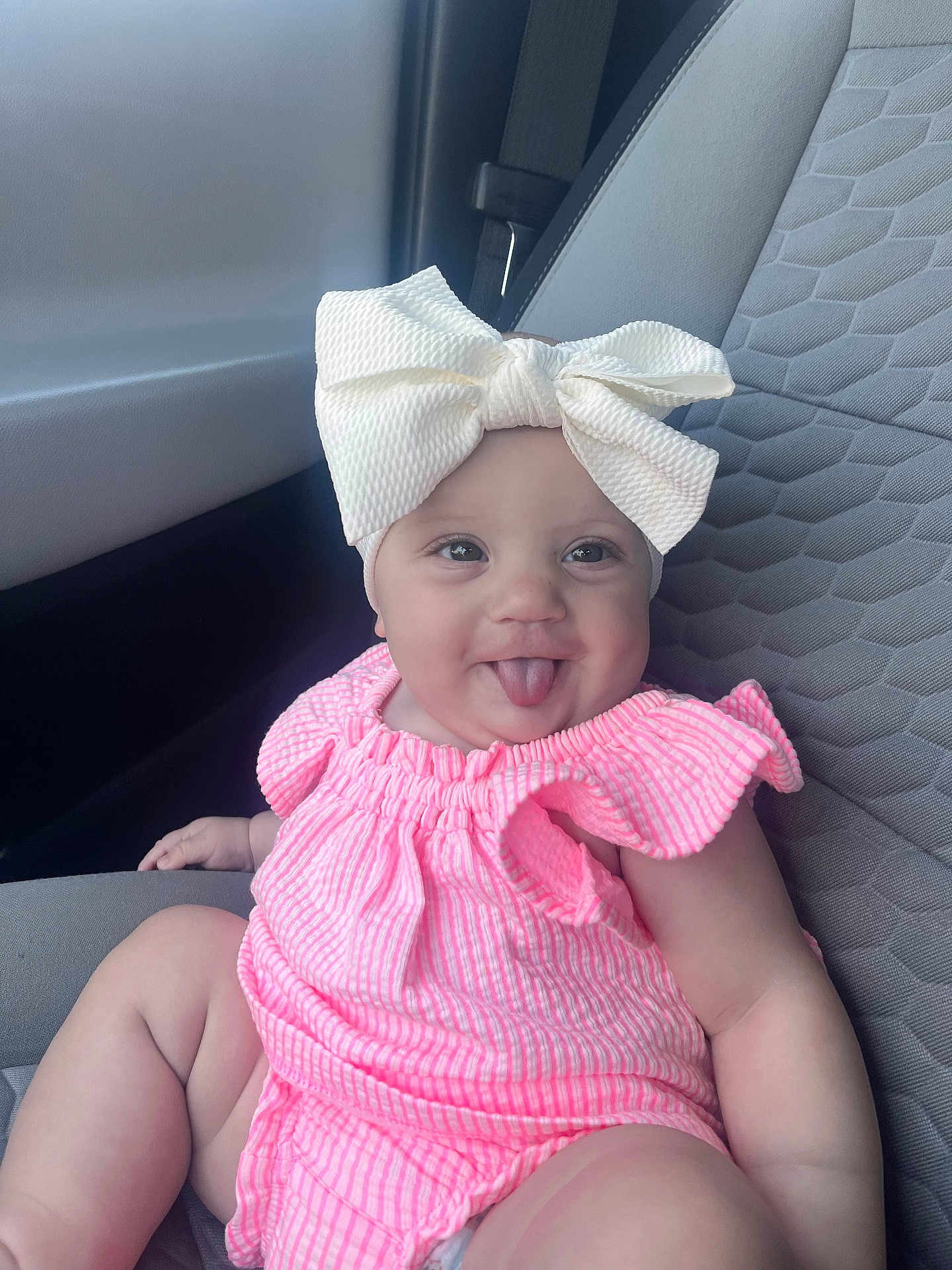 Kaislee is registered to the contest to win money with this photo: baby, child, tongue_out, white_bow, pink_outfit, car_seat, seatbelt, cute, smiling, chubby_arms, indoor, infant, clothing, headwear, person, portrait, happy, sitting, car_interior, seat