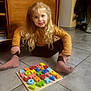 child, blonde_hair, sitting, floor, puzzle, alphabet, toy, colorful, socks, sweater, indoor, tiles, young, smiling, playtime, person, home, wooden_furniture, long_hair, casual_clothing