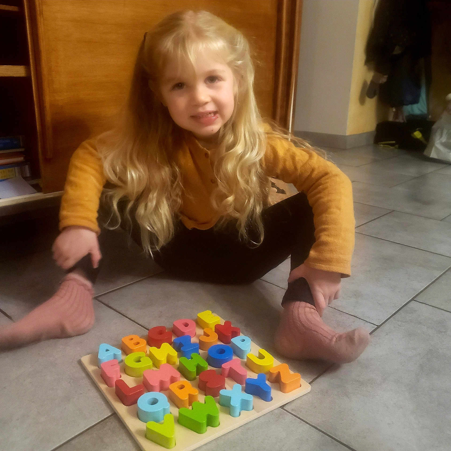 Sun a rejoint le concours — aidez-le/la à gagner de superbes lots ! alphabet, blonde_hair, casual_clothing, child, colorful, floor, home, indoor, long_hair, person, playtime, puzzle, sitting, smiling, socks, sweater, tiles, toy, wooden_furniture, young