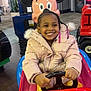 child, smiling, indoor, mall, kiddie_ride, car, steering_wheel, pink_jacket, braided_hair, beads, mickey_mouse, toy, playful, happy, portrait, colorful, fun, amusement, young_child, winter_clothing