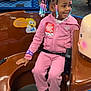 child, girl, smiling, amusement_ride, arcade, beads, braids, pink_clothing, shoes, indoor, playful, seatbelt, birthday_sticker, toy, game_machine, happy, colorful, entertainment, fun, leisure