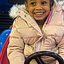 child, smiling, girl, braids, puffer_jacket, toy_car, steering_wheel, red, indoor, mickey_mouse, amusement, joy, winter_clothing, cute, portrait, happy, play, fun, kid, entertainment