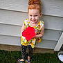 child, girl, redhead, hair_bun, smile, flower, red_flower, hibiscus, dress, sunflower_pattern, polka_dot_leggings, flip_flops, grass, backyard, house_siding, portrait, standing, happy, outdoor, cute