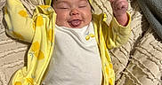 Emmarie Grace is registered to the contest to win money with this photo: baby, blanket, blue_pants, child, clothing, cute, expression, face, happy, headwear, indoors, infant, lying_down, person, raised_arms, smiling, texture, tongue_out, yellow_headband, yellow_jacket