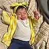 baby, blanket, blue_pants, child, clothing, cute, expression, face, happy, headwear, indoors, infant, lying_down, person, raised_arms, smiling, texture, tongue_out, yellow_headband, yellow_jacket