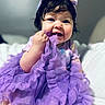 baby, child, girl, headband, purple_dress, fabric, chewing, smile, happy, cute, indoor, portrait, soft_lighting, chubby_cheeks, clothing, person, young_child, closeup, sitting, playful