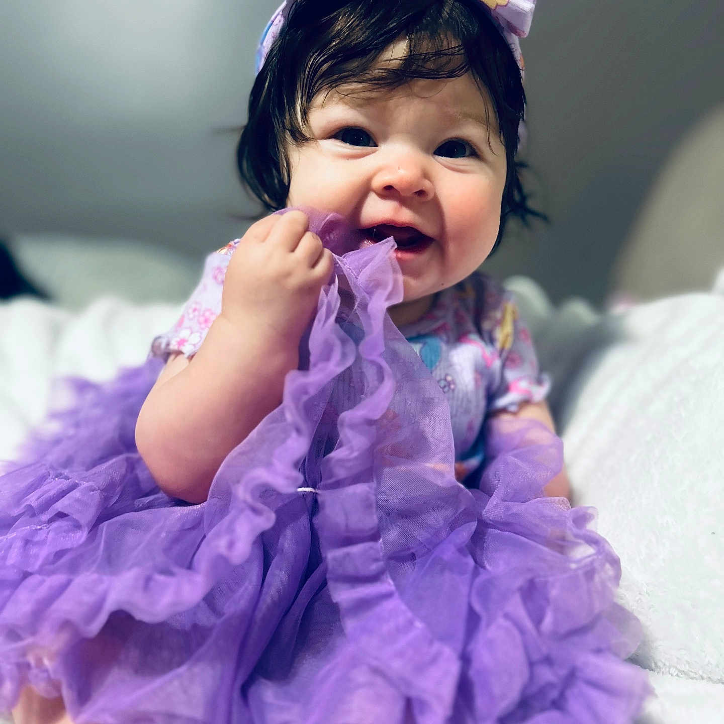Emmarie Grace is registered to the contest to win money with this photo: baby, child, girl, headband, purple_dress, fabric, chewing, smile, happy, cute, indoor, portrait, soft_lighting, chubby_cheeks, clothing, person, young_child, closeup, sitting, playful