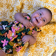Adeline is registered to the contest to win money with this photo: baby, blanket, child, clothing, colorful, cute, eyes, face, floral_dress, hand, happy, indoors, infant, lying_down, pattern, portrait, skin, smiling, yellow, young