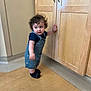 toddler, child, curly_hair, blue_outfit, standing, wooden_floor, cabinet, indoor, socks, bracelet, curious, person, young_child, home, furniture, looking_away, baby, flooring, light_skin, shorts