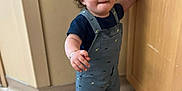 Rene joined the competition — help win amazing prizes! blue_clothing, bracelet, child, curly_hair, expression, floor, hand_on_cabinet, home, indoor, light_skin, overalls, person, room_corner, shorts, sock, standing, toddler, walking, wooden_cabinet, young_child
