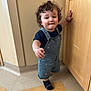 toddler, child, curly_hair, overalls, blue_clothing, indoor, wooden_cabinet, floor, sock, walking, hand_on_cabinet, expression, young_child, person, home, standing, light_skin, bracelet, shorts, room_corner
