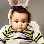 baby, child, toddler, curly_hair, bunny_ears, headband, striped_clothing, jacket, hoodie, hand, adult, indoor, portrait, cute, expression, face, person, sitting, gray_background, holding