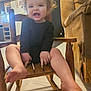 toddler, child, smiling, curly_hair, black_clothing, wooden_chair, rocking_chair, barefoot, indoor, living_room, couch, floor, happy, person, home, furniture, cute, portrait, baby, seated