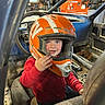 Jaxson joined the competition — help win amazing prizes! child, helmet, orange_helmet, red_jacket, vehicle, car_interior, steering_wheel, smiling, boy, workshop, tools, mechanic_shop, seat, window, face, hand, blue_eyes, dirt, metal, casual_clothing