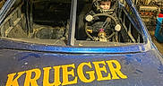 Jaxson is registered to the contest to win money with this photo: child, helmet, race_car, steering_wheel, garage, tools, machinery, advertisement, blue_car, yellow_text, dirt, gloves, young_driver, vehicle, indoor, racing, protective_gear, person, helmet_on_roof, workshop
