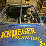 child, helmet, race_car, steering_wheel, garage, tools, machinery, advertisement, blue_car, yellow_text, dirt, gloves, young_driver, vehicle, indoor, racing, protective_gear, person, helmet_on_roof, workshop