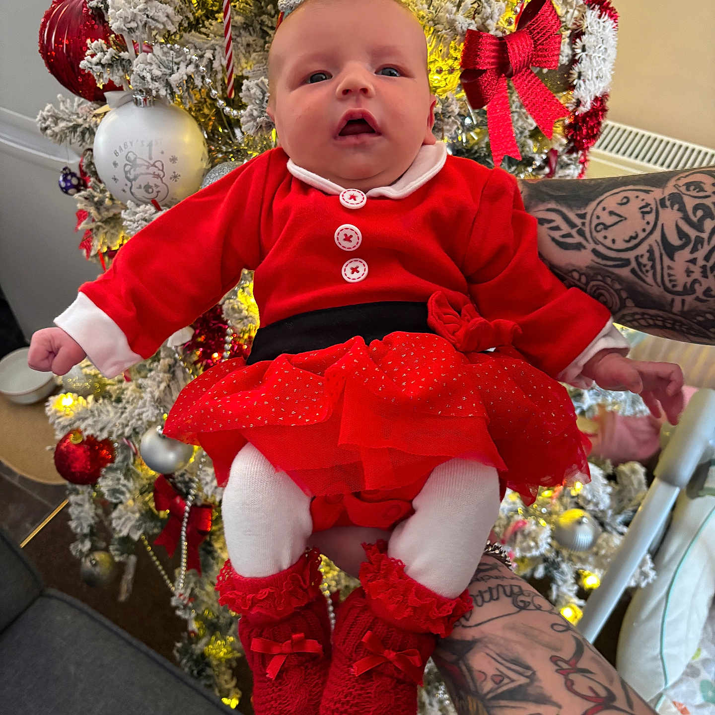 Nuala is registered to the contest to win money with this photo: baby, celebration, christmas_bow, christmas_tree, cute, decorations, festive, festivity, holding, holiday, holiday_season, indoors, infant, lights, ornaments, person, red_booties, red_clothing, tattoos, white_tights
