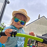 Esteban a rejoint le concours — aidez-le/la à gagner de superbes lots ! toddler, child, hat, sunglasses, bike_handlebar, outdoor, garden, toy, hand, smile, plaid_shirt, blue_shirt, green, playground, backyard, person, summer, casual_clothing, reflection, fun