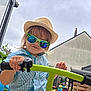 toddler, child, hat, sunglasses, bike_handlebar, outdoor, garden, toy, hand, smile, plaid_shirt, blue_shirt, green, playground, backyard, person, summer, casual_clothing, reflection, fun