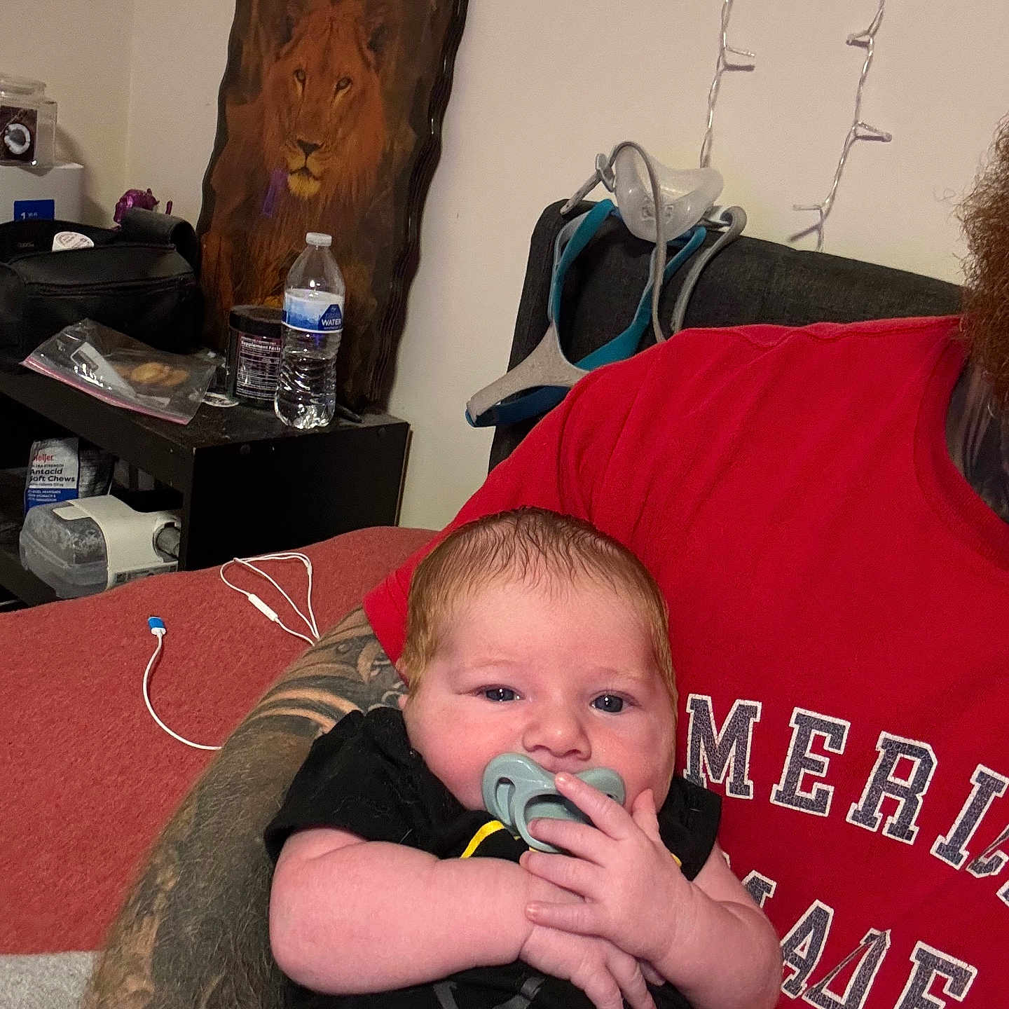 Sylys is registered to the contest to win money with this photo: arm, baby, casual, clothing, cute, face, furniture, hand, indoor, infant, lion_painting, newborn, pacifier, pacifier_in_mouth, person, portrait, red_shirt, resting, tattoo, water_bottle