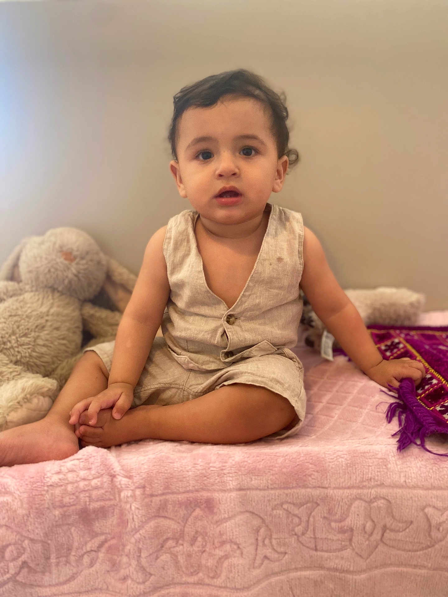 Adam a rejoint le concours — aidez-le/la à gagner de superbes lots ! toddler, child, baby, sitting, romper, beige_clothing, pink_blanket, plush_toy, bunny, indoor, curly_hair, hand_holding, purple_cloth, soft_lighting, wall, barefoot, cute, portrait, young_child, casual