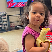 Celeste is registered to the contest to win money with this photo: toddler, child, banana, snack, indoor, living_room, television, purple_shirt, curly_hair, person, young_child, furniture, carpet, toy, hair_tie, face, candid, real_life, casual, home