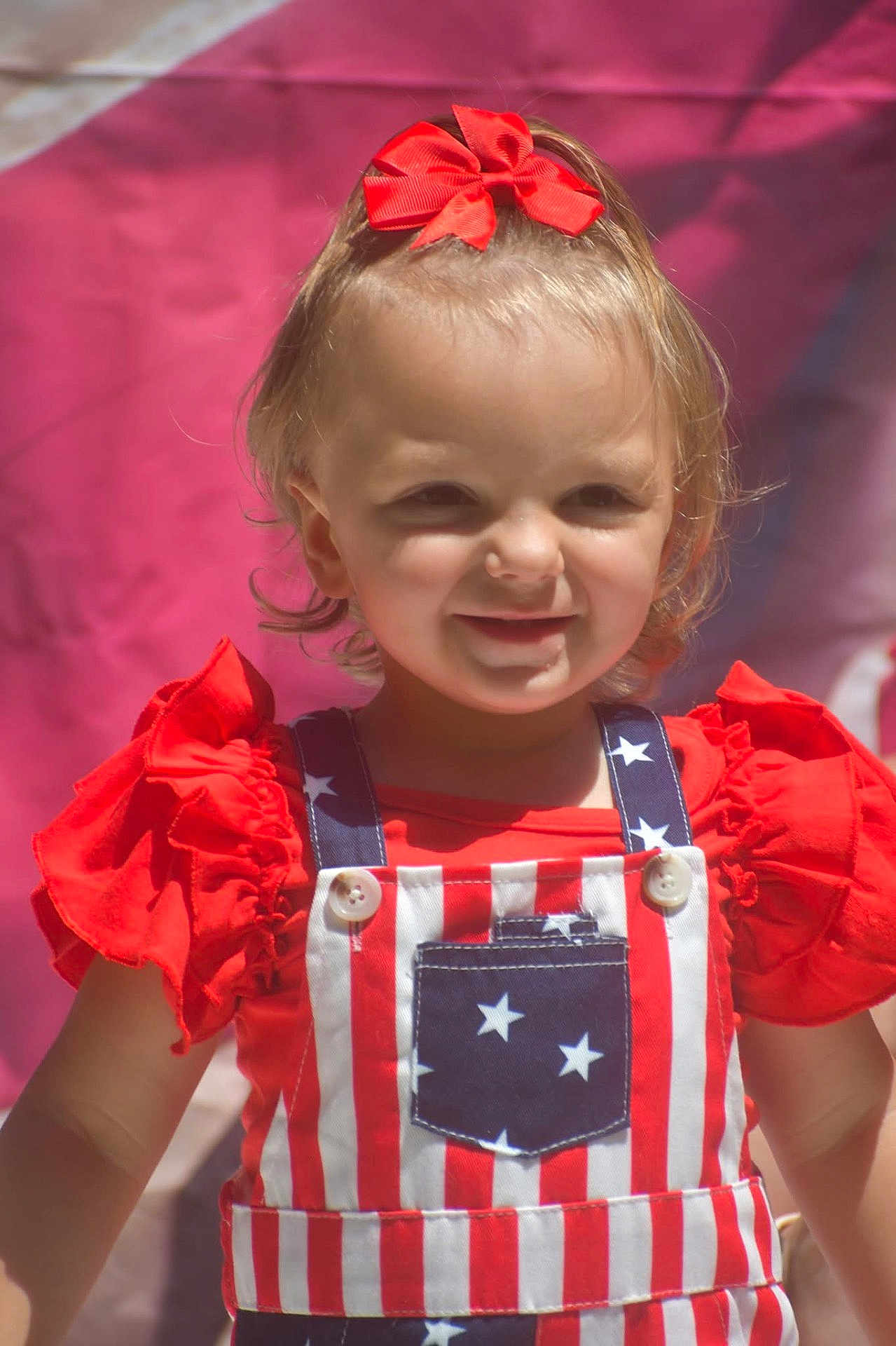 Emma joined the competition — help win amazing prizes! toddler, child, smiling, red_bow, overalls, stars, stripes, red_clothing, outdoor, portrait, happy, cute, baby, person, fashion, sunlight, face, head, background, colorful