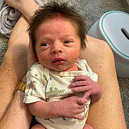 Luke is registered to the contest to win money with this photo: newborn, baby, infant, person, legs, freckles, hair, onesie, animal_print, hands, face, carpet, indoor, skin, adult, watch, stool, holding, cute, small
