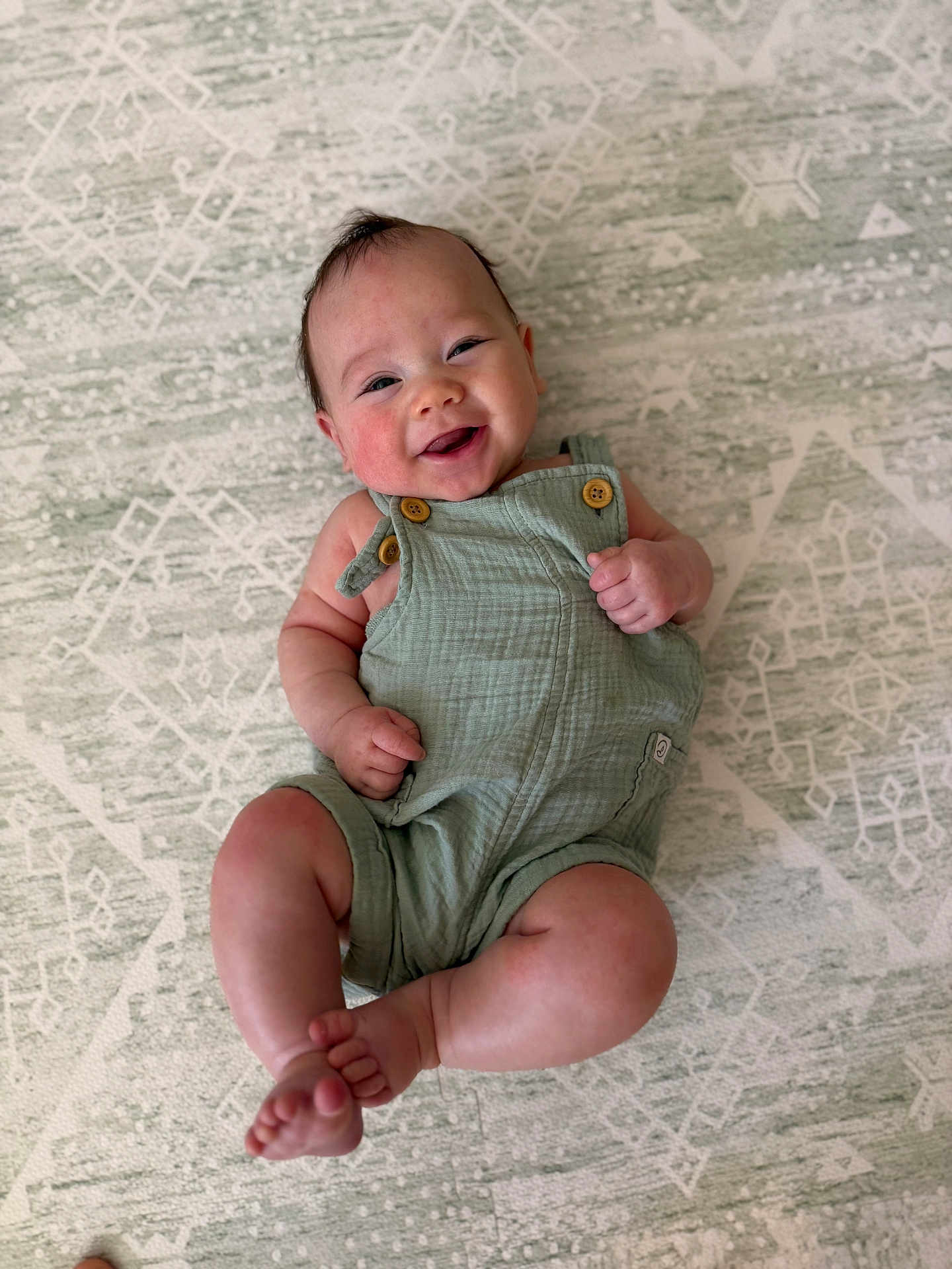 Easton joined the competition — help win amazing prizes! baby, infant, smiling, green_romper, patterned_rug, happy, lying_down, cute, chubby_legs, wooden_buttons, skin, head, arms, feet, toes, indoors, comfort, joy, portrait, child