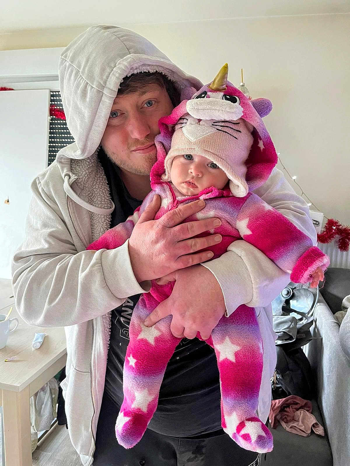 Eléna a rejoint le concours — aidez-le/la à gagner de superbes lots ! adult, baby, child, person, hoodie, unicorn_hat, onesie, pink_clothing, indoor, living_room, furniture, table, cup, hand, face, smile, cozy, casual, holding, curious