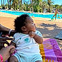 baby, child, poolside, swimming_pool, towel, summer, outdoor, palm_trees, water, playing, person, curly_hair, clothing, hand_holding, sunlight, relaxation, vacation, bright, colorful, daytime
