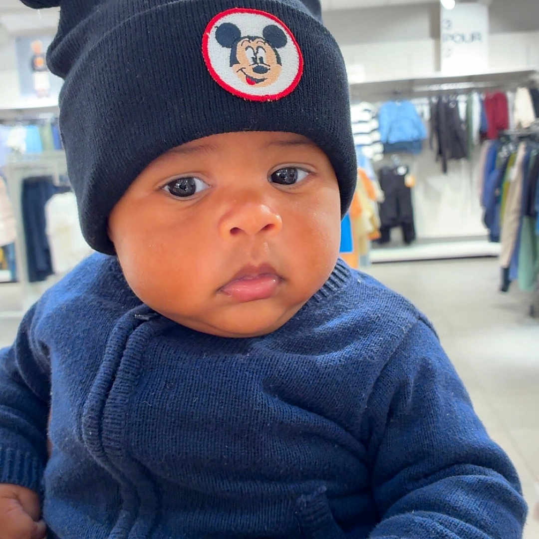 Izaric a rejoint le concours — aidez-le/la à gagner de superbes lots ! apparel, baby, black_hat, blue_sweater, child, clothing_store, cute, face, hand, headwear, indoor, infant, knitted_hat, mickey_mouse, person, portrait, serious_expression, shopping, store_rack, winter_clothing