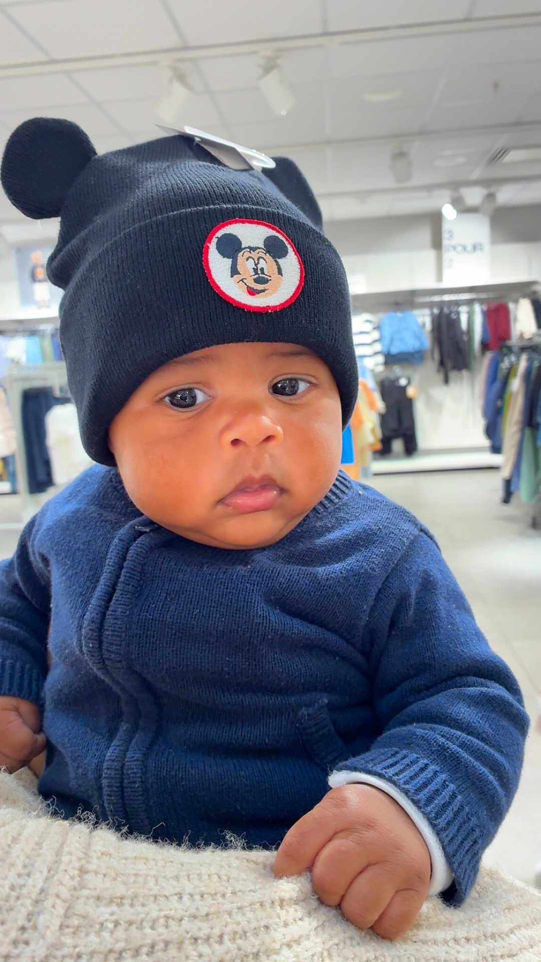 Izaric a rejoint le concours — aidez-le/la à gagner de superbes lots ! baby, child, blue_sweater, black_hat, mickey_mouse, indoor, clothing_store, face, headwear, cute, person, infant, portrait, hand, knitted_hat, winter_clothing, shopping, store_rack, apparel, serious_expression