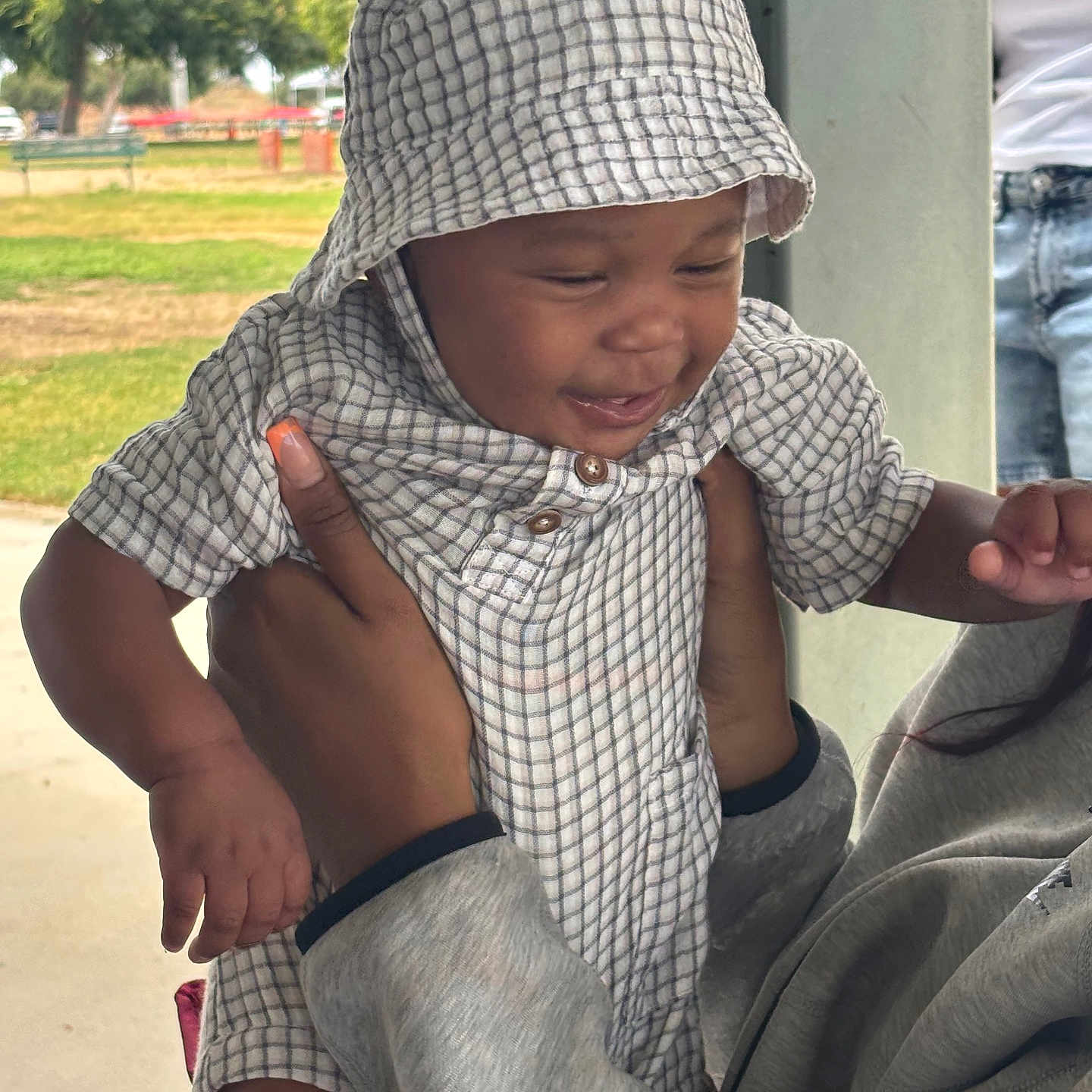 Masai Osho is registered to the contest to win money with this photo: baby, child, hood, checkered_clothing, smiling, hands, adult, outdoors, park, greenery, person, manicure, casual_wear, joyful, portrait, toddler, cute, holding, face, happy