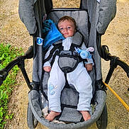 Jordy a rejoint le concours — aidez-le/la à gagner de superbes lots ! baby, stroller, child, onesie, plush_toy, blue, outdoor, grass, gravel, seat_belt, footwear, person, infant, cute, relaxed, daylight, nature, sitting, comfort, portrait