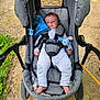 baby, stroller, child, onesie, plush_toy, blue, outdoor, grass, gravel, seat_belt, footwear, person, infant, cute, relaxed, daylight, nature, sitting, comfort, portrait