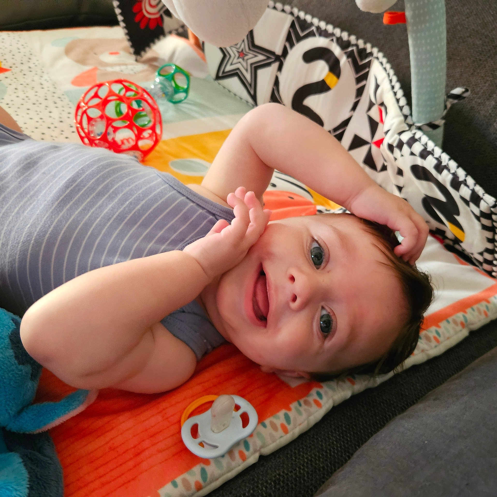 Jordy a rejoint le concours — aidez-le/la à gagner de superbes lots ! baby, blue_toy, child, closeup, clothing, colorful, cute, expression, face, hands, happy, indoor, infant, lying_down, pacifier, play_mat, playtime, smiling, soft, toy