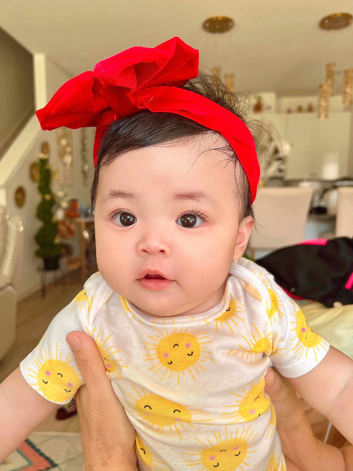Autumn Brynnah is registered to the contest to win money with this photo: baby, infant, child, red_headband, onesie, sun_pattern, cute, indoors, person, face, holding_hands, curious_eyes, soft_lighting, home_interior, furniture, smiling_sun, skin, hair, portrait, happy